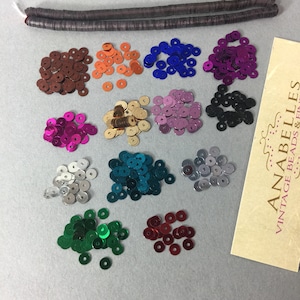 May include: Assortment of 12 colors of round sequins, each color in a separate pile. The sequins are metallic and have a shiny finish. The colors are: brown, orange, blue, purple, black, gold, pink, teal, silver, green, red, and white.  The sequins are arranged in two rows of six piles each.  The image also includes a brown, flat, rectangular piece of material with the text "ANABELLES VINTAGE BEADS &...