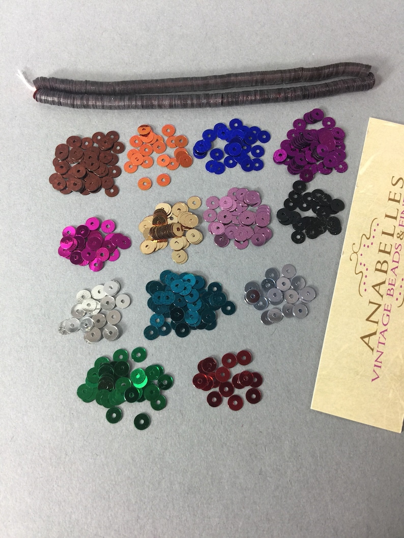 May include: Assortment of 12 colors of round sequins, each color in a separate pile. The sequins are metallic and have a shiny finish. The colors are: brown, orange, blue, purple, black, gold, pink, teal, silver, green, red, and white.  The sequins are arranged in two rows of six piles each.  The image also includes a brown, flat, rectangular piece of material with the text "ANABELLES VINTAGE BEADS &...