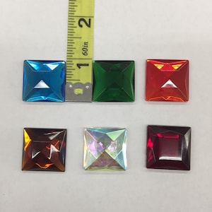 Puede incluir: Seis gemas cuadradas y facetadas de varios colores: azul, verde, rojo, marrón, iridiscente y rojo oscuro. Una cinta métrica amarilla muestra que las gemas miden aproximadamente 2,54 cm.