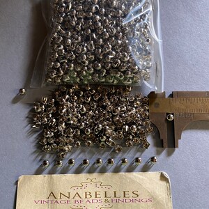 May include: A bag of silver metal rivets, with a few scattered on a table. The rivets are approximately 1.2 cm in diameter. A business card for Anabelles Vintage Beads & Findings is also on the table.