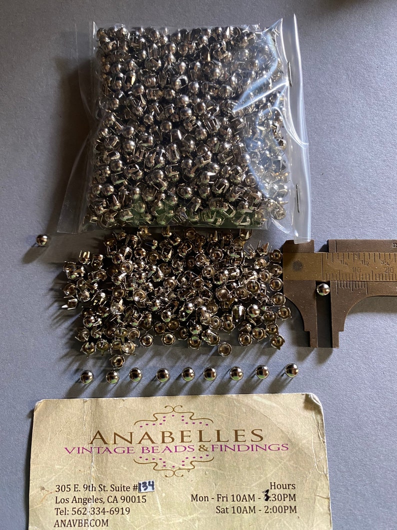 May include: A bag of silver metal rivets, with a few scattered on a table. The rivets are approximately 1.2 cm in diameter. A business card for Anabelles Vintage Beads & Findings is also on the table.