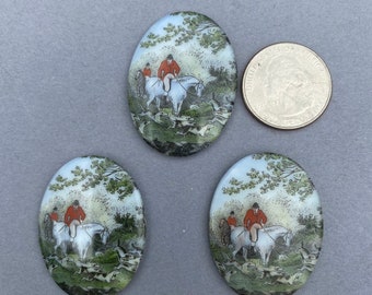 May include: Three oval-shaped glass cabochons with a painted scene of a fox hunt. The scene features riders on horseback with hounds running through a wooded area. The cabochons are approximately 1 inch in diameter.