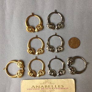 May include: A collection of vintage metal jewelry findings, including gold-toned and silver-toned ram head pendants. The pendants are circular with a ram head design on each side. A business card is also present.
