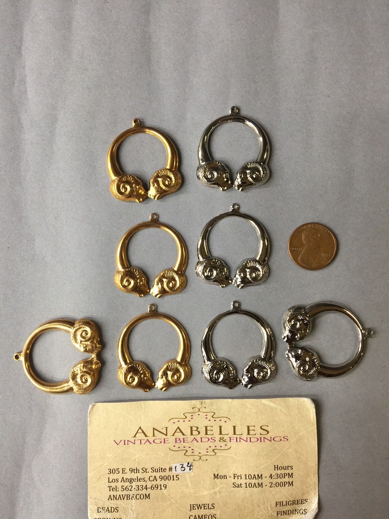 May include: A collection of vintage metal jewelry findings, including gold-toned and silver-toned ram head pendants. The pendants are circular with a ram head design on each side. A business card is also present.