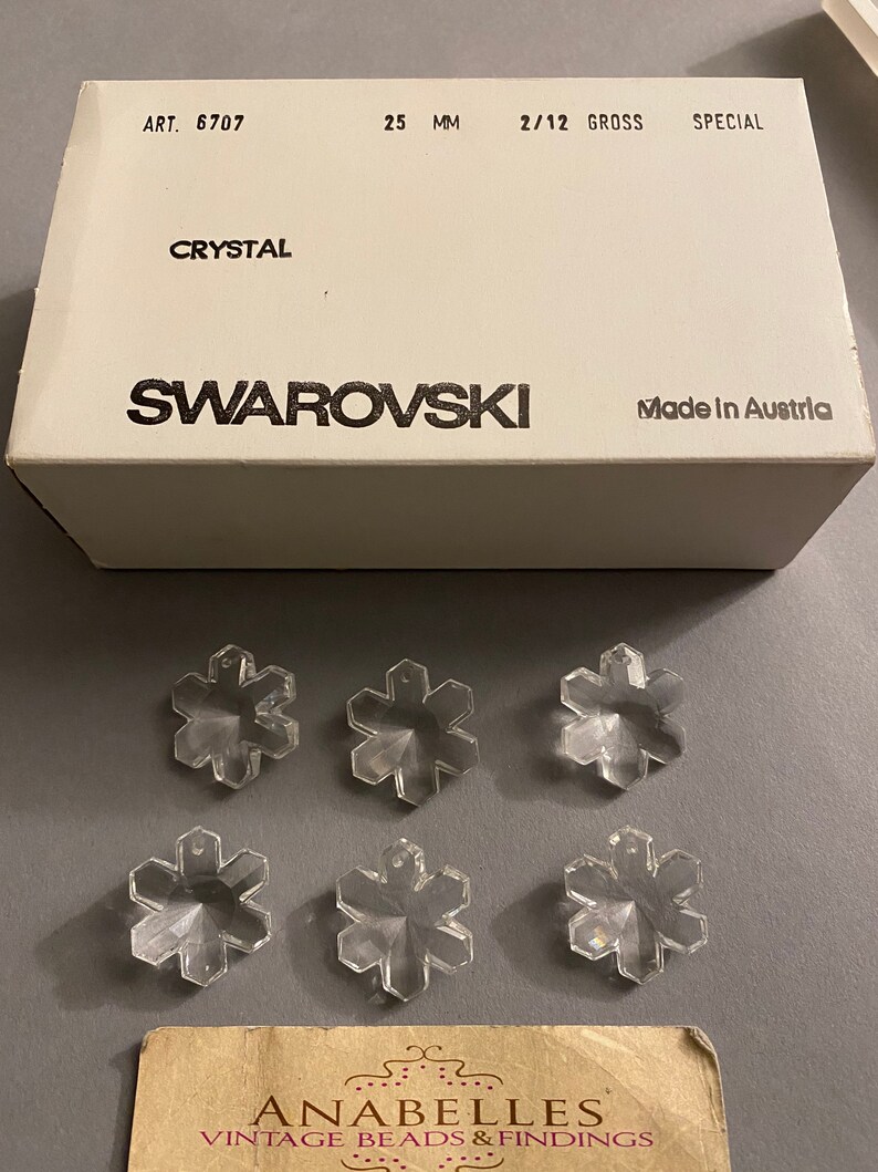 May include: A box of clear crystal snowflake charms, labeled "Swarovski" and "Made in Austria". The box also includes the text "ART. 6707", "25 MM", "2/12 GROSS", and "SPECIAL".