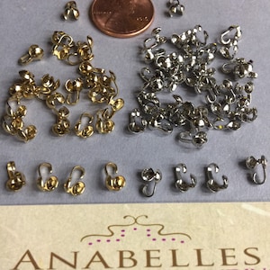 May include: A collection of gold and silver metal bead caps. The caps are small and have a rounded top with a loop on each side. The caps are arranged in a pile on a white surface. A US penny is included for scale.