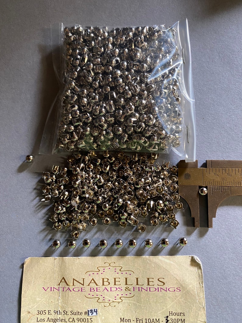May include: A bag of silver metal rivets, with a ruler measuring the size of the rivets. The rivets are approximately 1.2 cm in diameter. The bag is labelled "Anabelles Vintage Beads & Findings" with the address "305 E. 9th St. Suite #134 Los Angeles, CA 90015" and the hours "Mon - Fri 10AM - 3:30PM".