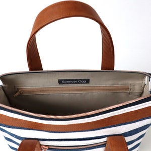 May include: Inside view of a striped navy blue and white canvas tote bag with brown leather handles and trim. The bag has a zippered pocket and a label that reads "Spencer Ogg".