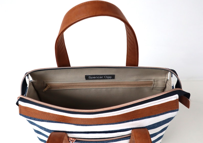 May include: Inside view of a striped navy blue and white canvas tote bag with brown leather handles and trim. The bag has a zippered pocket and a label that reads "Spencer Ogg".