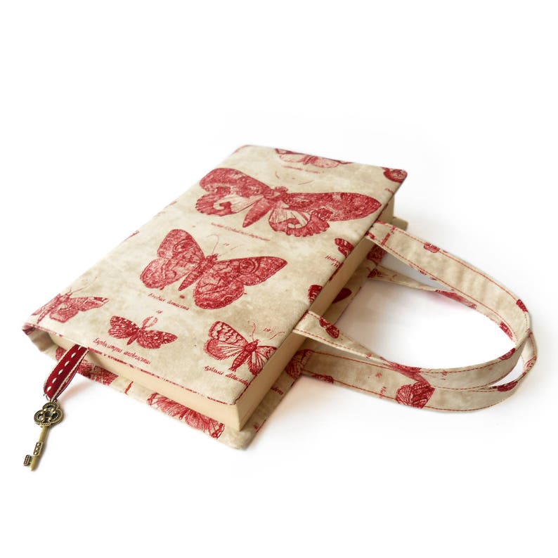 May include: A beige book cover with red butterfly illustrations and a key charm attached to a red ribbon. The cover has two fabric handles.
