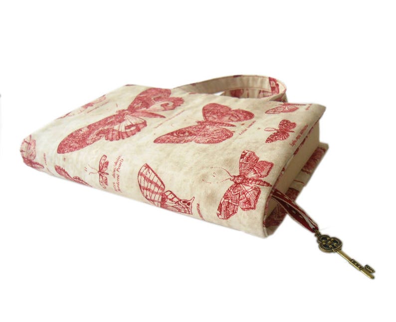 May include: A beige book cover with a red butterfly design. The cover has a red ribbon bookmark with a gold key charm.