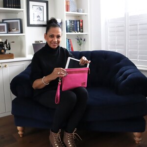May include: A woman is sitting on a blue velvet sofa holding a pink leather tablet case with a tablet inside. The case has a wrist strap.