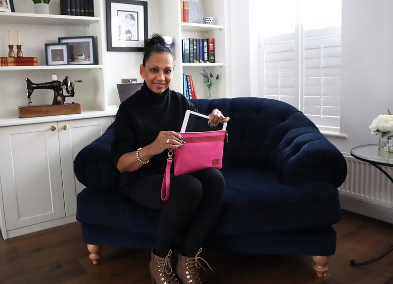 May include: A woman is sitting on a blue velvet sofa holding a pink leather tablet case with a tablet inside. The case has a wrist strap.