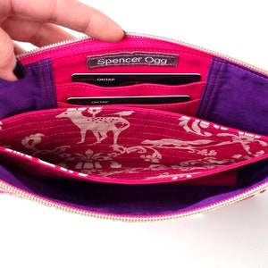 May include: Open pink fabric clutch with a purple lining and multiple card slots. The clutch has a gold zipper and a gold chain strap. The interior fabric has a white deer print on a pink background.