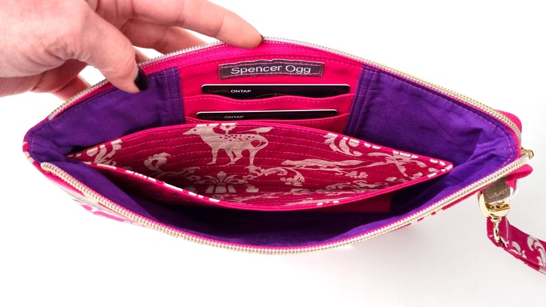 May include: Open pink fabric clutch with a purple lining and multiple card slots. The clutch has a gold zipper and a gold chain strap. The interior fabric has a white deer print on a pink background.