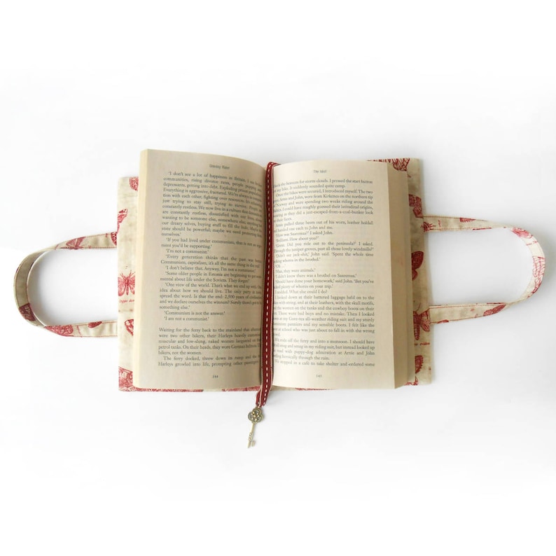May include: A book cover with a red and white butterfly pattern. The cover is made of fabric and has a red ribbon bookmark. The book is open to a page with text. The book is sitting on a white surface.