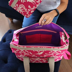 May include: A pink tote bag with a white floral pattern and a purple lining. The bag has two pockets inside and a zipper closure. The bag has two handles.
