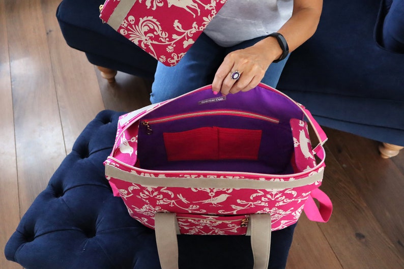 May include: A pink tote bag with a white floral pattern and a purple lining. The bag has two pockets inside and a zipper closure. The bag has two handles.