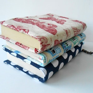 May include: A stack of four books with fabric covers. The top book has a red and white butterfly pattern, the second book has a blue and white house pattern, the third book has a blue and white polka dot pattern, and the bottom book is white.