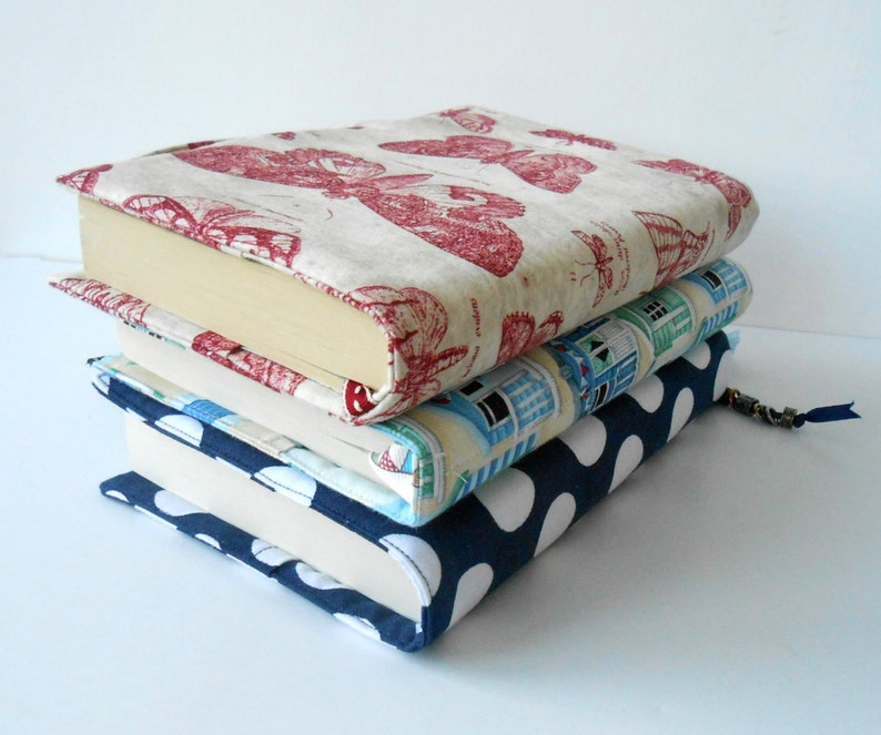 May include: A stack of four books with fabric covers. The top book has a red and white butterfly pattern, the second book has a blue and white house pattern, the third book has a blue and white polka dot pattern, and the bottom book is white.