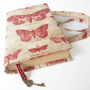 May include: A beige book cover with red butterfly illustrations and a key charm. The cover has two fabric handles.