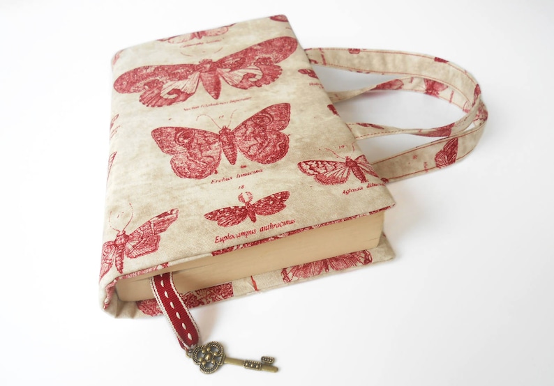 May include: A beige book cover with red butterfly illustrations and a key charm. The cover has two fabric handles.