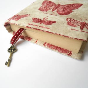 May include: A book cover with a beige background and red butterfly illustrations. The cover has a red ribbon bookmark with a decorative key charm.