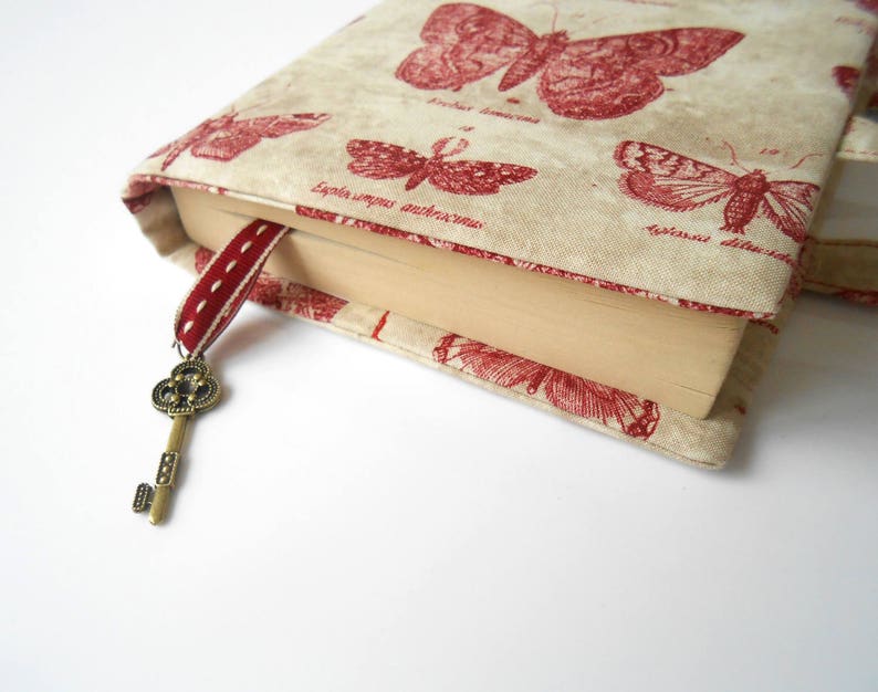 May include: A book cover with a beige background and red butterfly illustrations. The cover has a red ribbon bookmark with a decorative key charm.