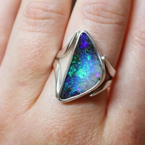 K&ouml;nnte beinhalten: Silberring mit einem dreieckigen Opal in leuchtenden Blau-, Gr&uuml;n- und Lilat&ouml;nen. Der Opal ist in einem schlichten Silberband gefasst, das die nat&uuml;rliche Sch&ouml;nheit des Edelsteins hervorhebt. Ein auff&auml;lliges Schmuckst&uuml;ck.