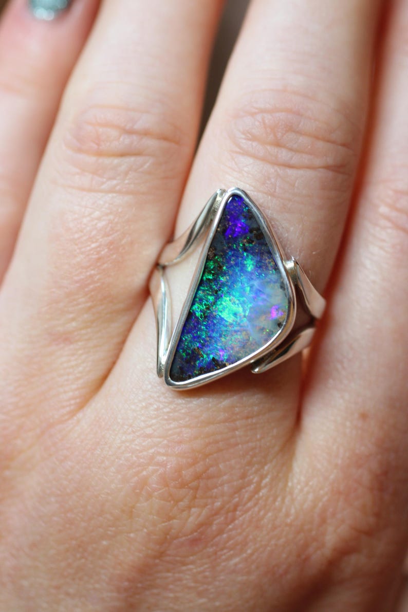 K&ouml;nnte beinhalten: Silberring mit einem dreieckigen Opal in leuchtenden Blau-, Gr&uuml;n- und Lilat&ouml;nen. Der Opal ist in einem schlichten Silberband gefasst, das die nat&uuml;rliche Sch&ouml;nheit des Edelsteins hervorhebt. Ein auff&auml;lliges Schmuckst&uuml;ck.