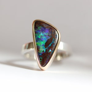 Könnte beinhalten: Ein silberner Ring mit einem dreieckigen Opal-Edelstein, der in einer goldenen Fassung sitzt. Der Opal hat eine dunkelbraune Basis mit grün, blau und violetten Schimmern.