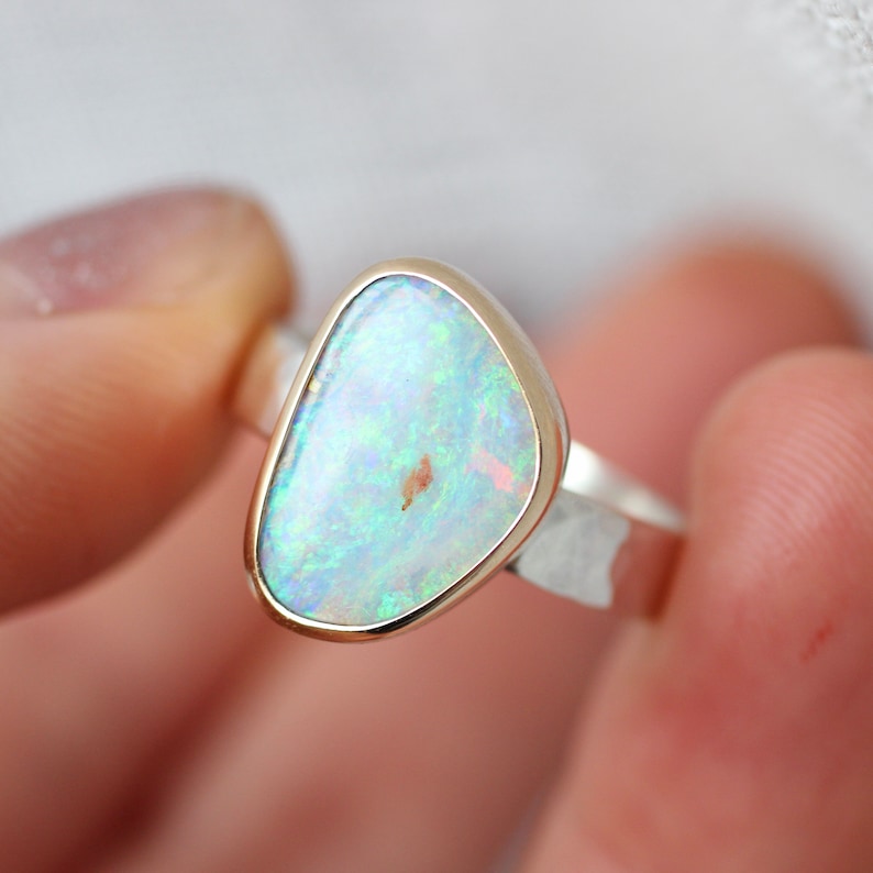 May include: A silver ring with a white opal gemstone set in a gold bezel. The opal has a light blue and green iridescent sheen.