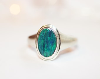 May include: A silver ring with an oval-shaped, iridescent green and blue opal stone. The opal is set in a silver bezel, and the band is smooth and polished. The ring is displayed on a white surface.