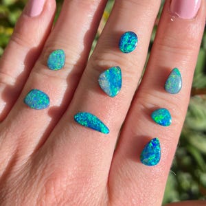 Op de afbeelding: Een verzameling van zeven blauw-groene opalen in verschillende vormen en maten. De opalen worden getoond op een hand, waardoor hun iriserende kleuren worden getoond.