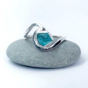 Può includere: Un anello d'argento con un design a spirale presenta una pietra preziosa blu chiaro. L'anello è posto su una pietra grigia.
