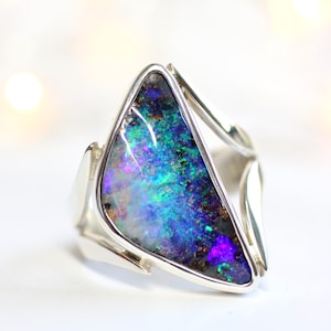 K&ouml;nnte beinhalten: Silberring mit einem dreieckigen Opalstein. Der Opal zeigt eine lebendige Farbpalette, darunter Blau-, Gr&uuml;n- und Lilat&ouml;ne. Der Ring hat ein modernes, offenes Banddesign.