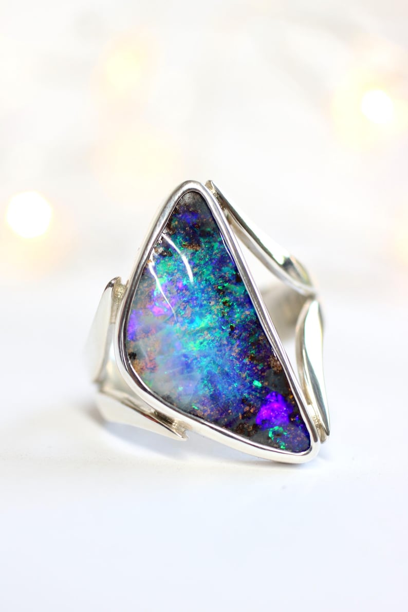 K&ouml;nnte beinhalten: Silberring mit einem dreieckigen Opalstein. Der Opal zeigt eine lebendige Farbpalette, darunter Blau-, Gr&uuml;n- und Lilat&ouml;ne. Der Ring hat ein modernes, offenes Banddesign.