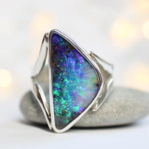 K&ouml;nnte beinhalten: Ein Silberring mit einem gro&szlig;en, schillernden Opal, der lebendige Blau-, Gr&uuml;n- und Lilat&ouml;ne aufweist. Der Opal ist in einem geometrischen, modernen Design gefasst und liegt auf einem grauen Stein vor unscharfem Hintergrund.