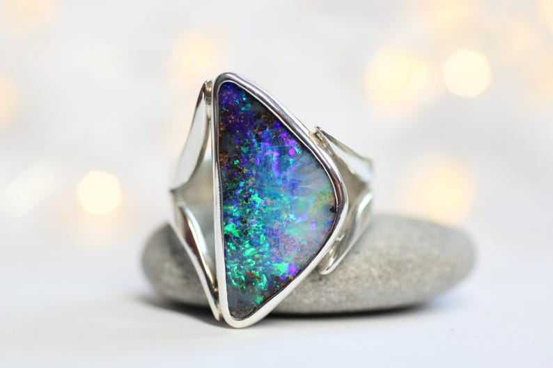 K&ouml;nnte beinhalten: Ein Silberring mit einem gro&szlig;en, schillernden Opal, der lebendige Blau-, Gr&uuml;n- und Lilat&ouml;ne aufweist. Der Opal ist in einem geometrischen, modernen Design gefasst und liegt auf einem grauen Stein vor unscharfem Hintergrund.