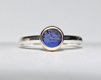 May include: A silver ring with a round, deep blue stone set in a gold bezel. The ring has a simple, elegant design, with a thin silver band. The blue stone has a textured appearance.