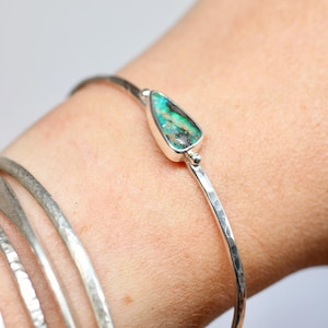 May include: A silver bangle bracelet featuring a teardrop-shaped turquoise stone with dark matrix patterns. The metal band has a hammered, textured finish. The bracelet is worn alongside three additional plain, hammered silver bangles.