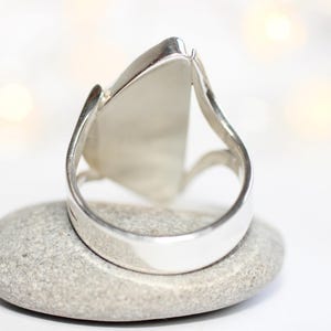 K&ouml;nnte beinhalten: Ein silberner Ring mit einem gro&szlig;en, dreieckigen, hellfarbigen Stein. Der Ring liegt auf einem kleinen, grauen Stein. Der Ring hat ein dickes, glattes Band mit einem geschwungenen Design auf beiden Seiten des Steins.