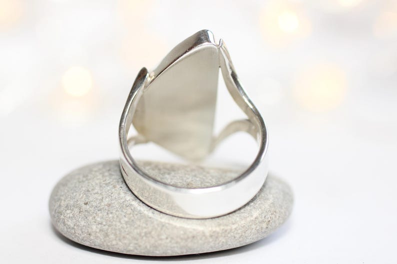 K&ouml;nnte beinhalten: Ein silberner Ring mit einem gro&szlig;en, dreieckigen, hellfarbigen Stein. Der Ring liegt auf einem kleinen, grauen Stein. Der Ring hat ein dickes, glattes Band mit einem geschwungenen Design auf beiden Seiten des Steins.