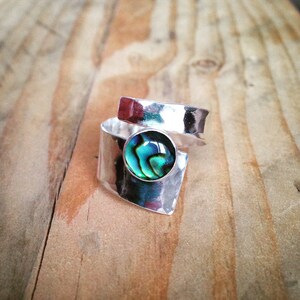 Può includere: Due anelli d'argento. Uno presenta una grande conchiglia di abalone rotonda e iridescente. L'altro è una semplice fascia. Gli anelli sono su uno sfondo di legno. La conchiglia di abalone ha sfumature di blu, verde e nero.