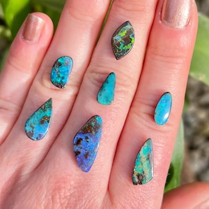 May include: A collection of seven polished opal gemstones in various shapes and sizes. The opals display vibrant colors, including blues, greens, and teals, with dark matrix patterns. The gemstones are held in a hand, showcasing their natural beauty.