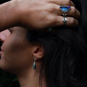 K&ouml;nnte beinhalten: Ein silberner Ring mit einem dreieckigen, schillernden Stein und ein kleinerer Silberring an einem Finger. Ein silberner Ohrring mit einem T&uuml;rkisstein ist sichtbar. Der Schmuck vor dunklem Hintergrund.