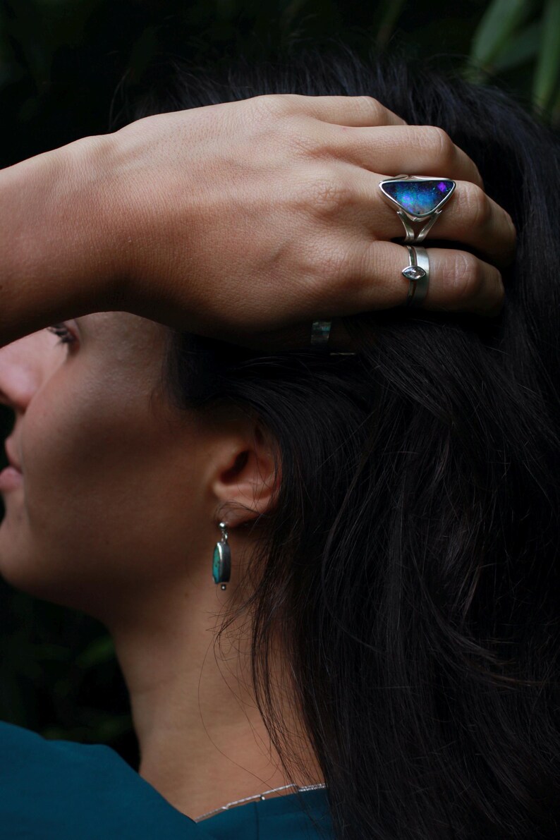 K&ouml;nnte beinhalten: Ein silberner Ring mit einem dreieckigen, schillernden Stein und ein kleinerer Silberring an einem Finger. Ein silberner Ohrring mit einem T&uuml;rkisstein ist sichtbar. Der Schmuck vor dunklem Hintergrund.