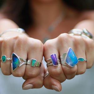 K&ouml;nnte beinhalten: Nahaufnahme von H&auml;nden, die mehrere Silberringe mit farbenfrohen, schillernden Edelsteinen zeigen. Die Ringe weisen Steine in verschiedenen Formen und Gr&ouml;&szlig;en in Blau-, Gr&uuml;n- und Lilat&ouml;nen auf, die in Silberb&auml;ndern gefasst sind. Der Hintergrund ist unscharf.