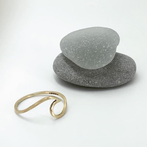 Pode incluir: Um anel de ouro em forma de onda sobre uma superfície branca com duas pedras cinzas empilhadas uma em cima da outra.