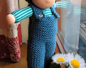 May include: A knitted doll wearing a burgundy knit hat with a pom pom and blue overalls. The doll is standing in front of a window with a white curtain and a reflection of the doll in the glass. The doll is looking out the window at a white daisy.